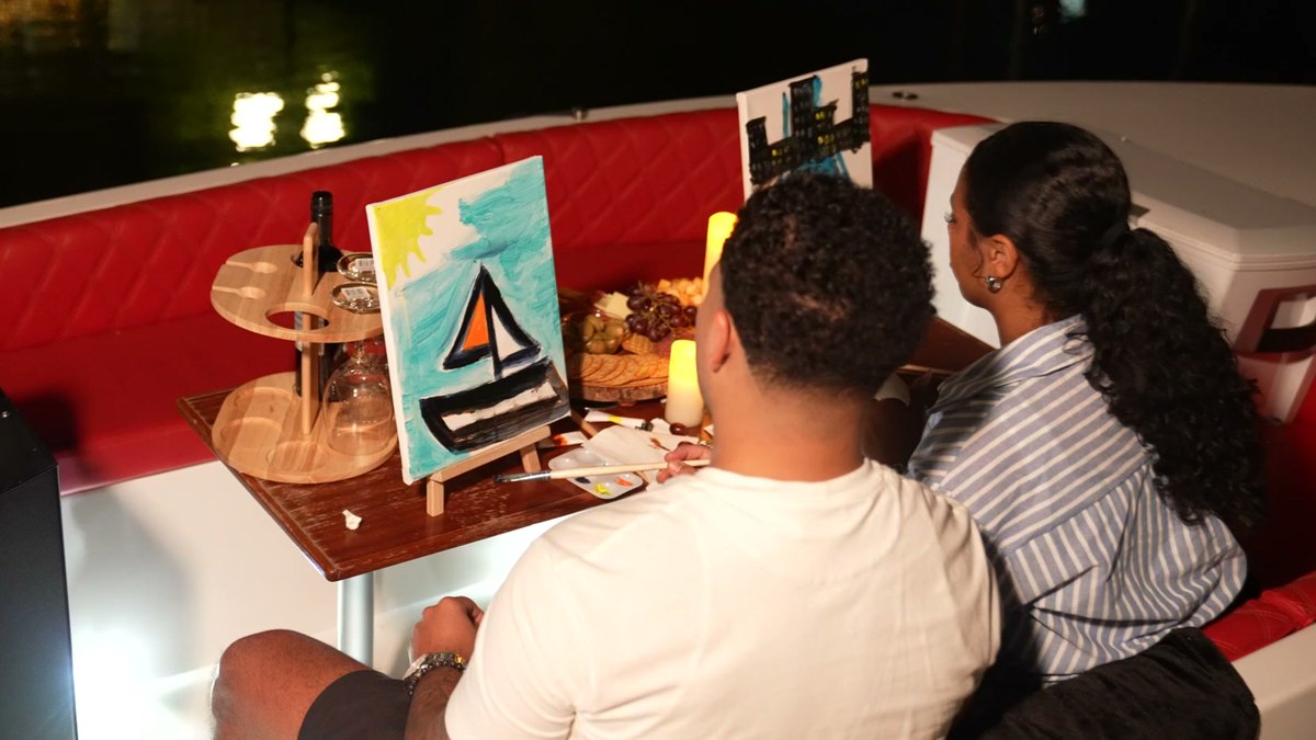 A couple on a private paint-and-sip date night aboard a Yachtsy boat, painting sailboats on easels by candlelight