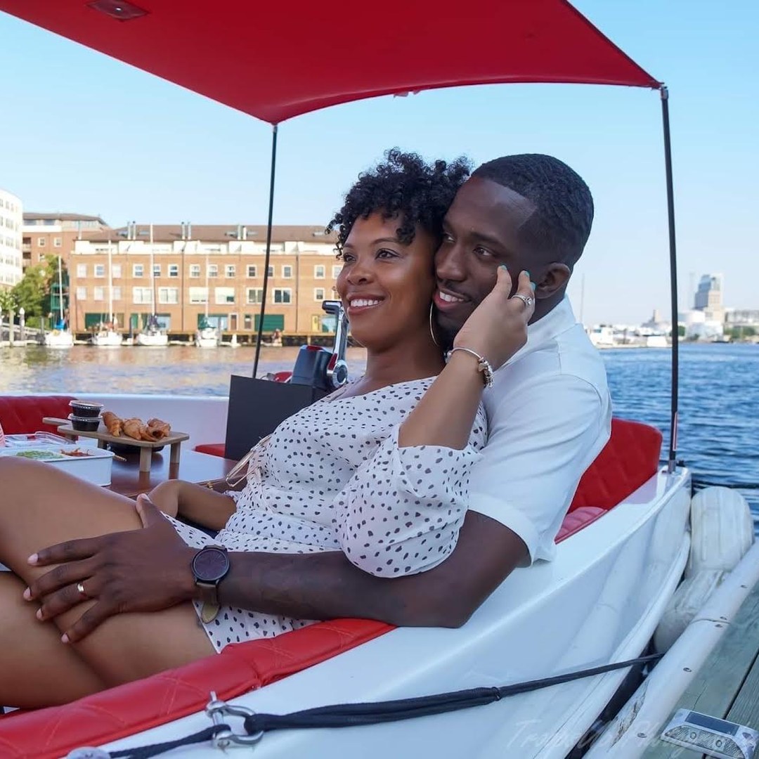 A couple sitting close together under the red canopy of a private Yachtsy picnic boat at golden hour, the harbor skyline and a picnic grazing board behind them